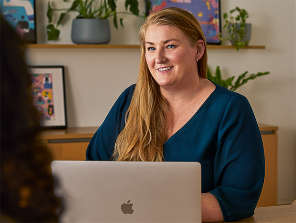 Een Apple medewerker zit in een gezamenlijke ruimte aan een tafel achter een MacBook en praat met een collega.