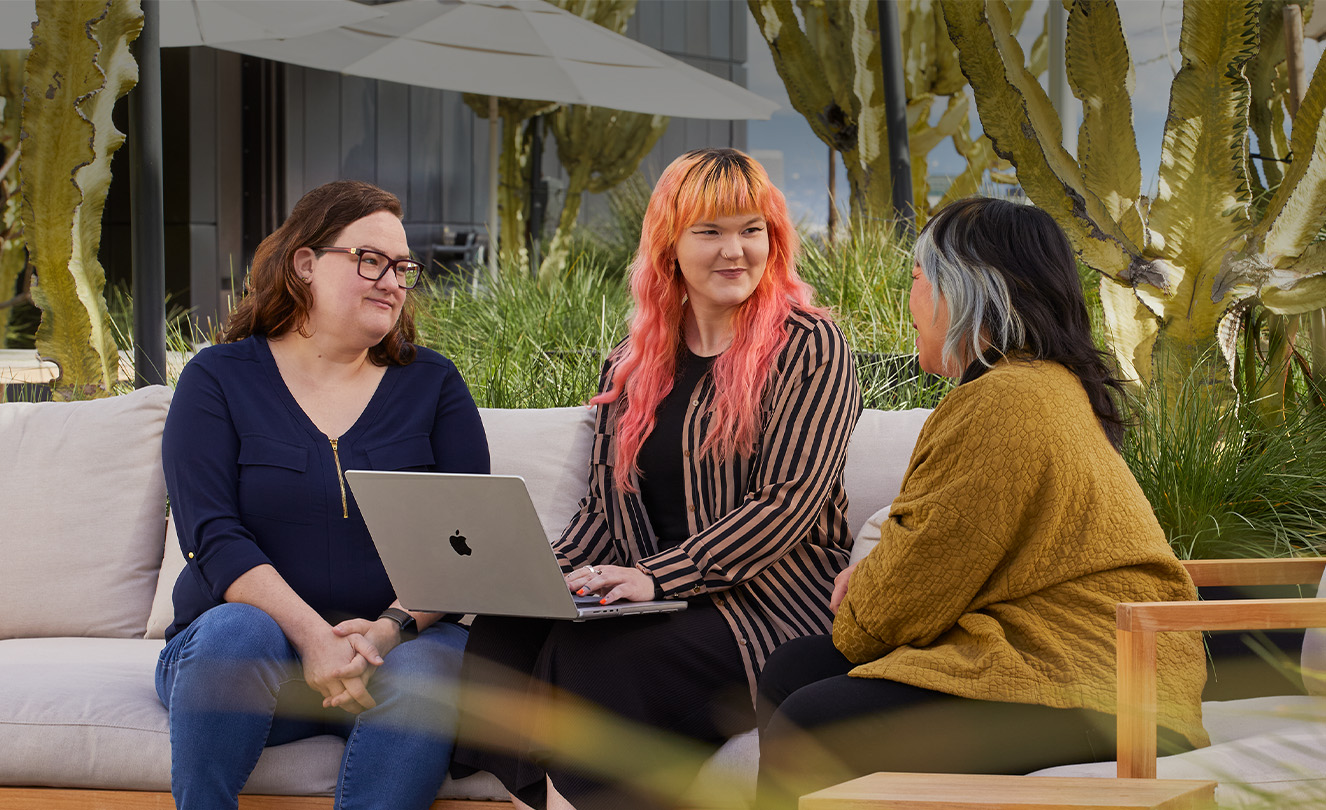 Drie Apple medewerkers, waarvan een met een MacBook, zitten samen op een terras.