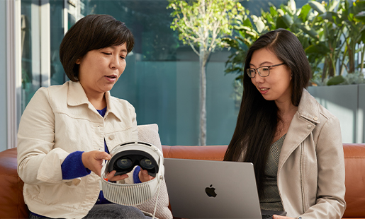 Jessica zit binnen op een MacBook te werken. Ze zit naast een open deur naar het terras, samen met een Apple collega die haar de Apple Vision Pro laat zien.
