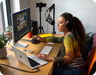 امرأة في منزلها تعمل خلف مكتبها على MacBook Pro متصل بشاشتين خارجيتين وترتدي سماعات رأس وتتحدث في الميكروفون