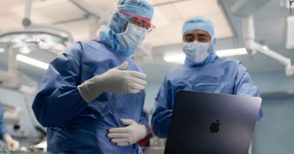 Två kirurger står i en operationssal och tittar på en MacBook. Den ena gestikulerar med handen medan den andra ser på.