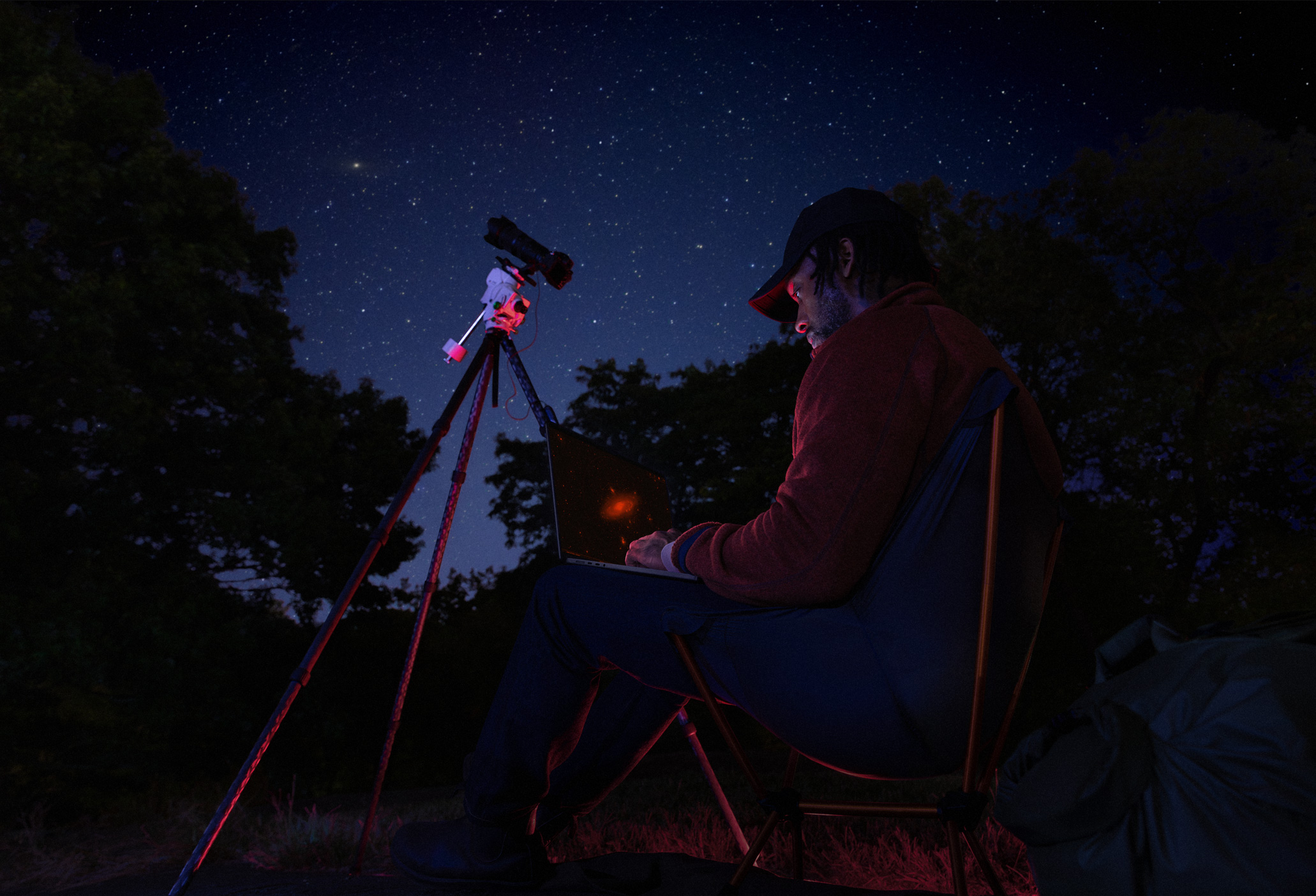Personne assise dehors la nuit avec un MacBook Pro sur les genoux, connecté à un appareil sur un trépied qui pointe vers le ciel étoilé
